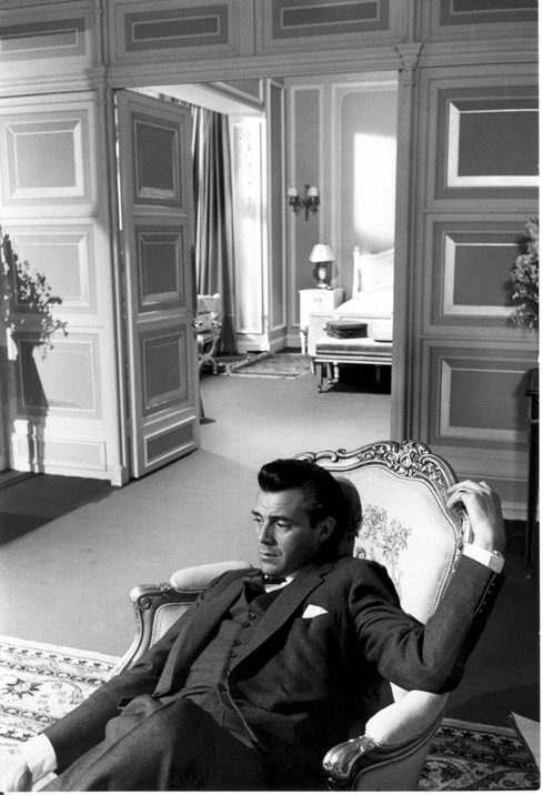 A black and white photo of actor Dir Bogarde. He's a handsome white man with dark hair, dark eyes, lounging in a chair wearing a smart suit. He's in the lower right of the photo with a posh hotel room in the background